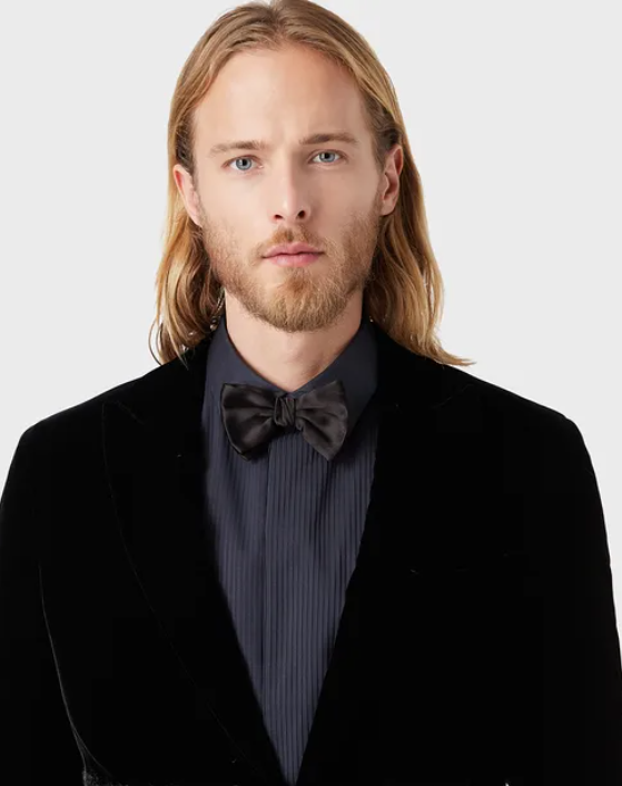 A young man with shoulder length hair wears a dark blue button up shirt, black blazer and bow tie.